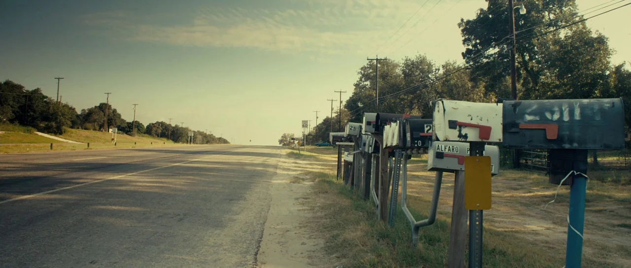 The Imposter (2012) movie still: establishing — A rural asphalt road extends toward the horizon under a clear daytime sky. A series of weathered…