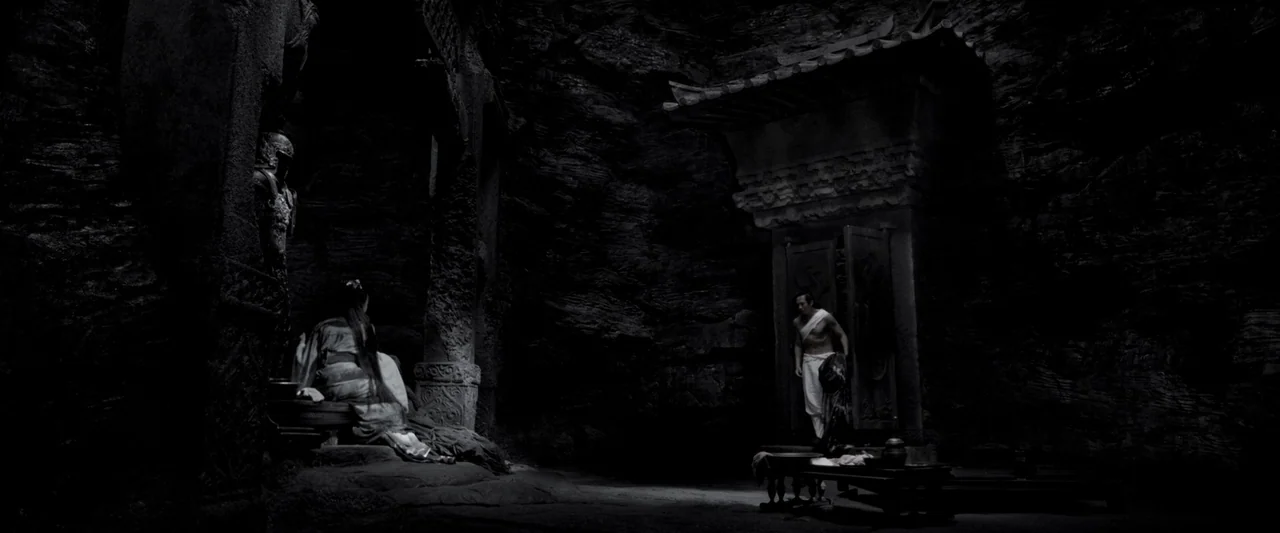 Shadow (2018) movie still: wide — A dark, cavernous space with raw stone walls and carved structural pillars. A shirtless man stands in a carved…