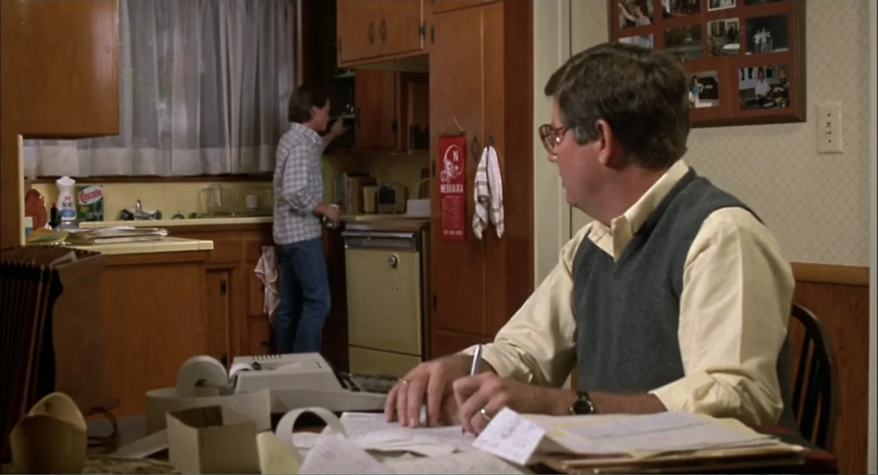 Teen Wolf (1985) movie still: medium — A 1980s residential home interior with wood cabinetry and a cluttered dining table. A man works at a table as…