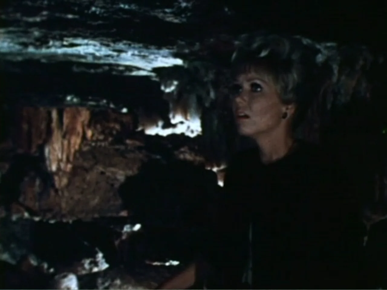 It's Alive (1974) movie still: medium — A dark subterranean cave with textured rock walls and cavernous shadows. A woman with blonde hair stands…