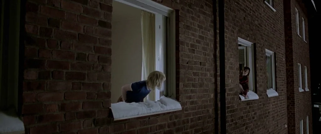 Let The Right One In (2008) movie still: wide — A dark red brick exterior of an apartment building features two illuminated window openings. Two…
