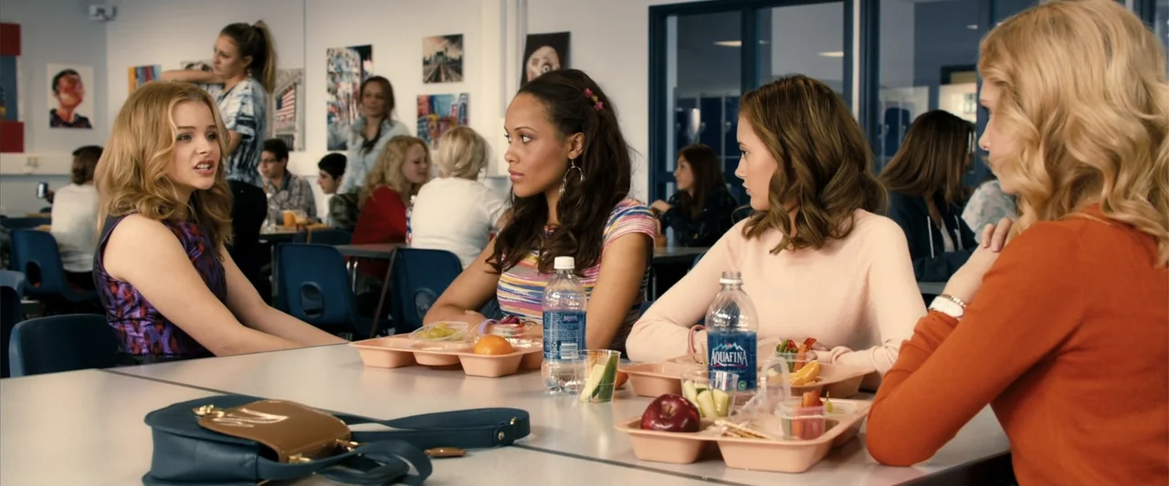 Kick-Ass 2 (2013) movie still: medium — A high school lunch room with white walls, blue chairs, and scattered students. Four teenage girls are…