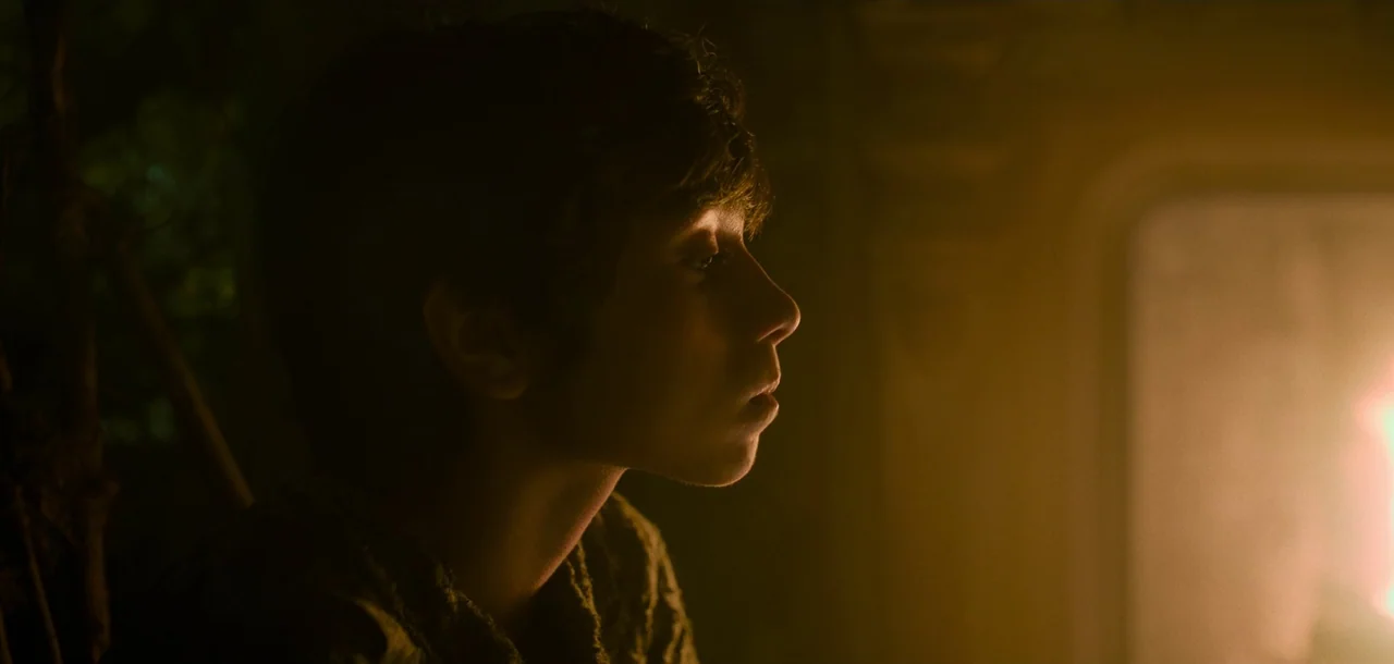 Peter Pan & Wendy (2023) movie still: closeup — A dimly lit interior space is dominated by the warm glow of a fireplace. A teenager with wavy hair…
