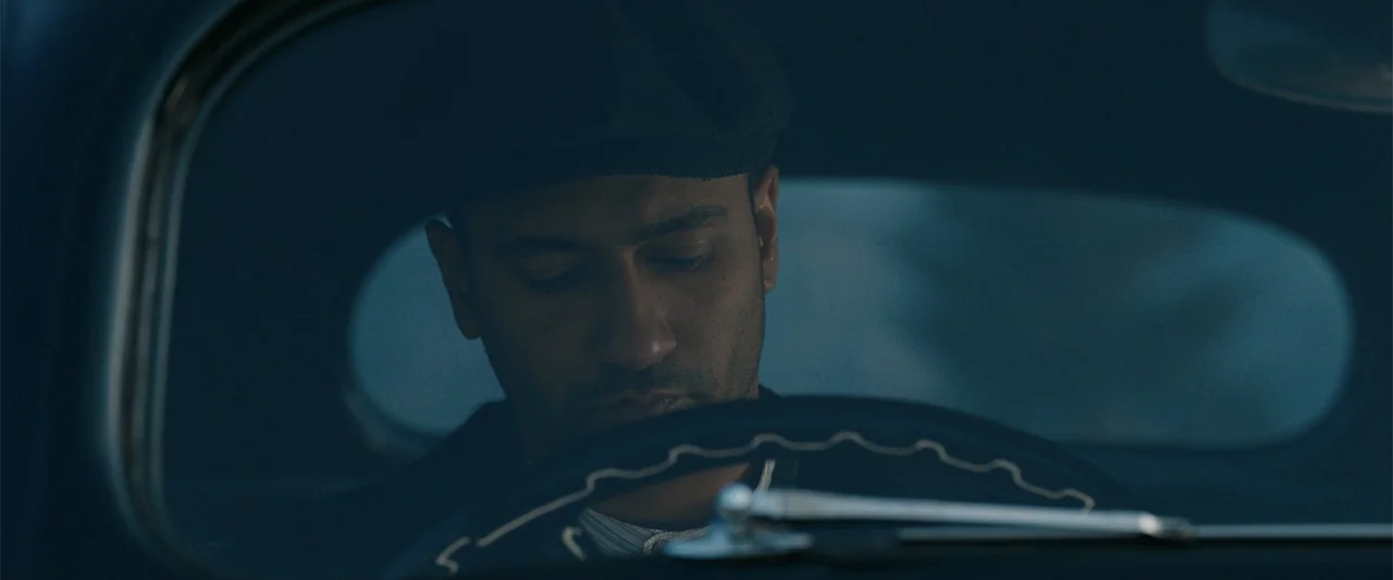 Sardar Udham (2021) movie still: closeup — The interior of a vintage automobile is dark and shadowed. A man wearing a cap sits behind the wheel,…