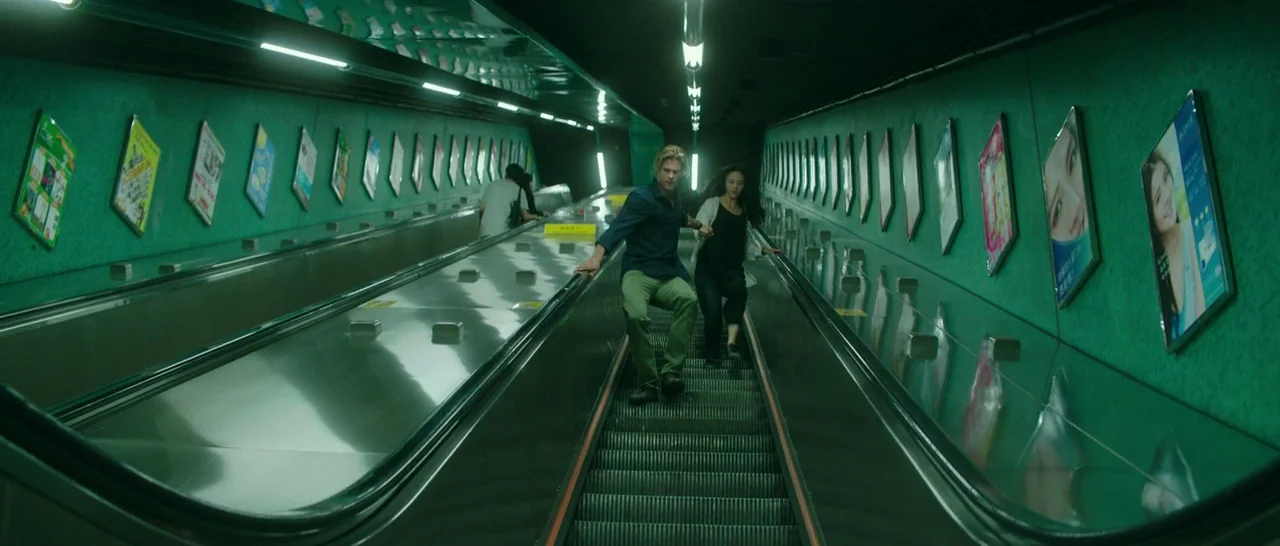 Blackhat (2015) movie still: fullbody — A long escalator descends through a tunnel lined with vibrant teal walls and advertisements. A man and a…
