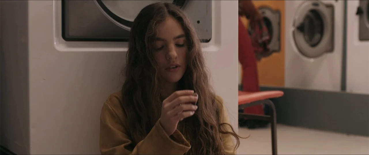 Woman of the Hour (2024) movie still: medium — A modern, sterile laundromat with rows of white washing machines and a red plastic seat. A teenage…