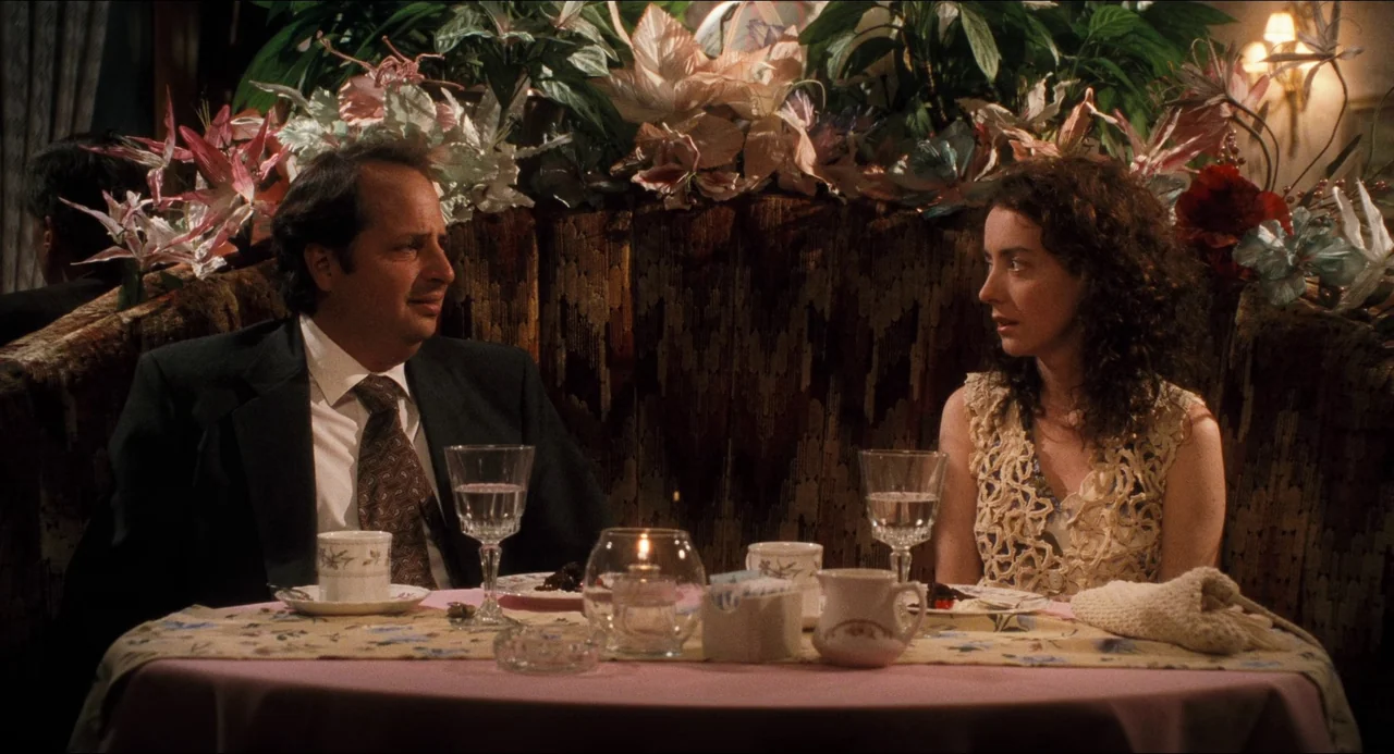 Happiness (1998) movie still: medium — A dark restaurant booth features a pink-clothed table covered in dinnerware and floral arrangements. A man in…
