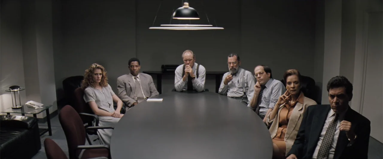 The Pelican Brief (1993) movie still: wide — A sparse conference room features a large black oval table centered under hanging lights. Seven…