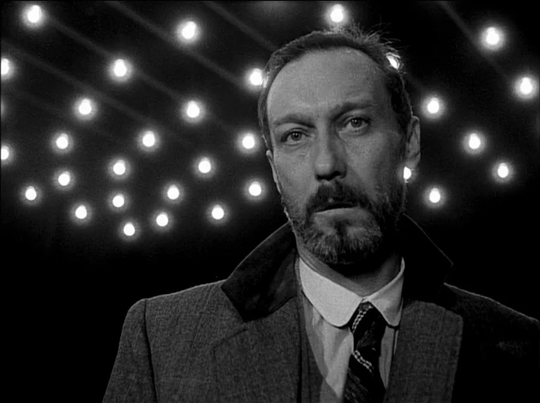 My 20th Century (1989) movie still: closeup — A man stands before a grid of exposed light bulbs in a dark space. He looks toward the camera while…