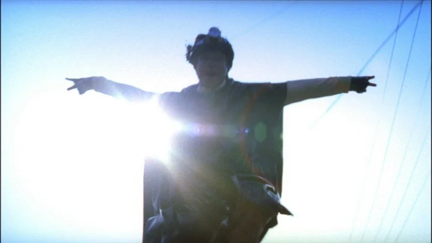 Save The Green Planet! (2003) movie still: medium — An open outdoor area against a clear blue sky during the day. A person wearing a cape and…