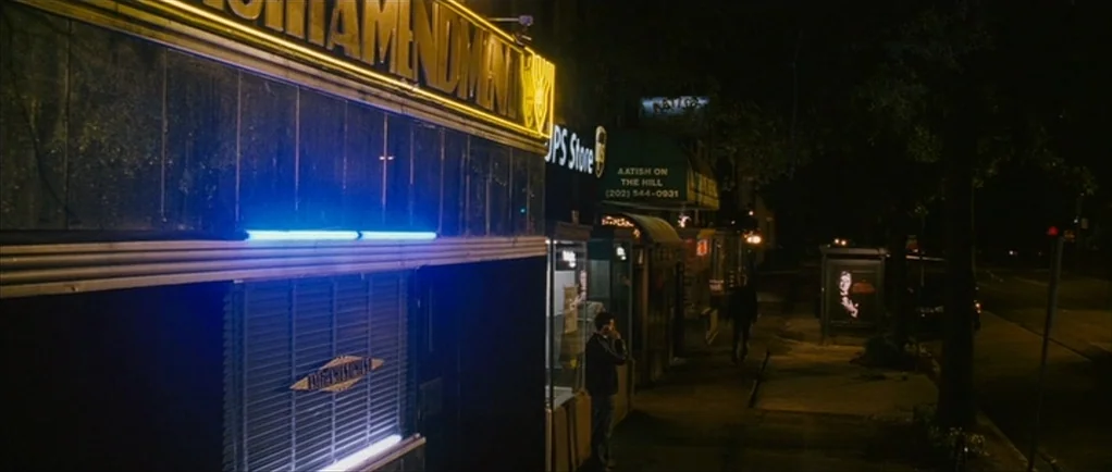 The Walker (2007) movie still: wide — A quiet urban sidewalk at night features lit business storefronts under a dark sky. A bright yellow neon sign…