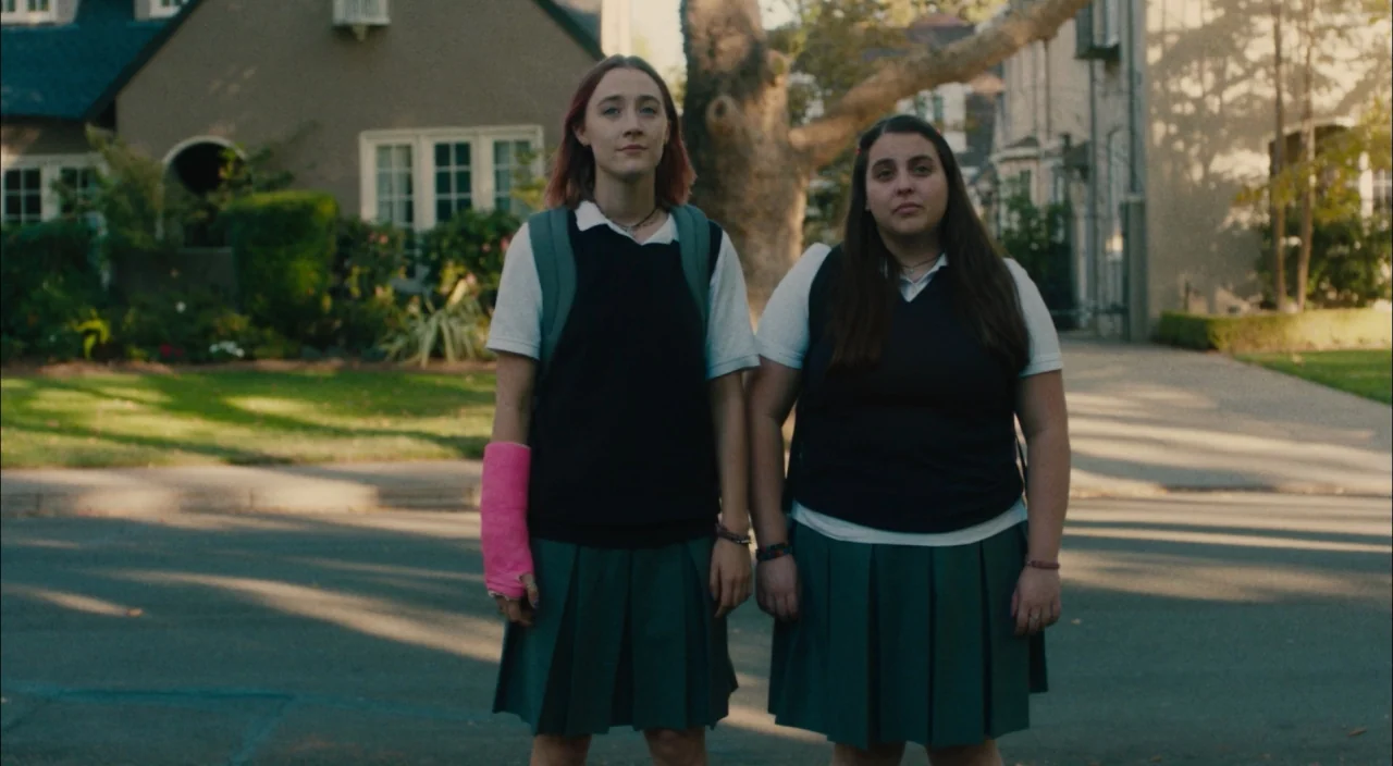 Lady Bird (2017) movie still: fullbody — Two teenage girls stand on a residential asphalt street in front of a house. The girls are wearing matching…