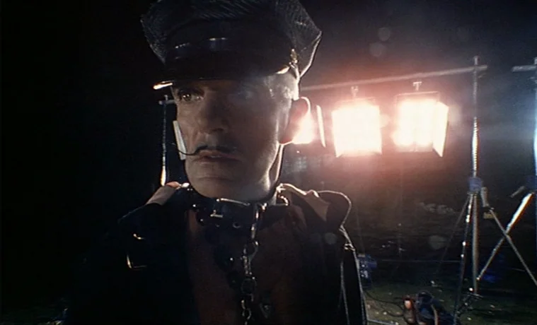The Garden (1990) movie still: medium — A dark outdoor space contains illuminated production lighting equipment on metal stands. An individual with…