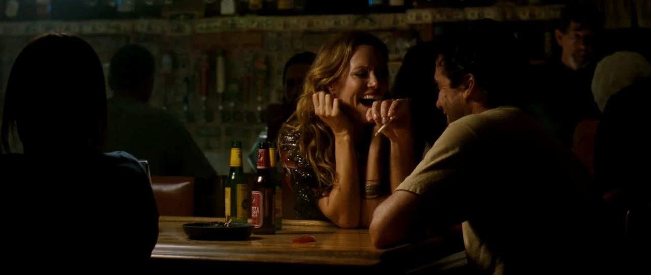 Little Birds (2011) movie still: medium — A dimly lit bar setting with beer bottles and an ashtray on the table. A man and a woman sit facing each…