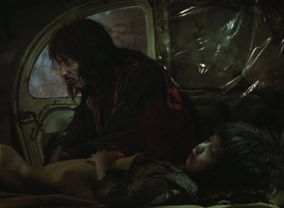Dodes’ka-den (1970) movie still: medium — The interior of a derelict car with plastic sheeting covering the broken windows. A disheveled man watches…