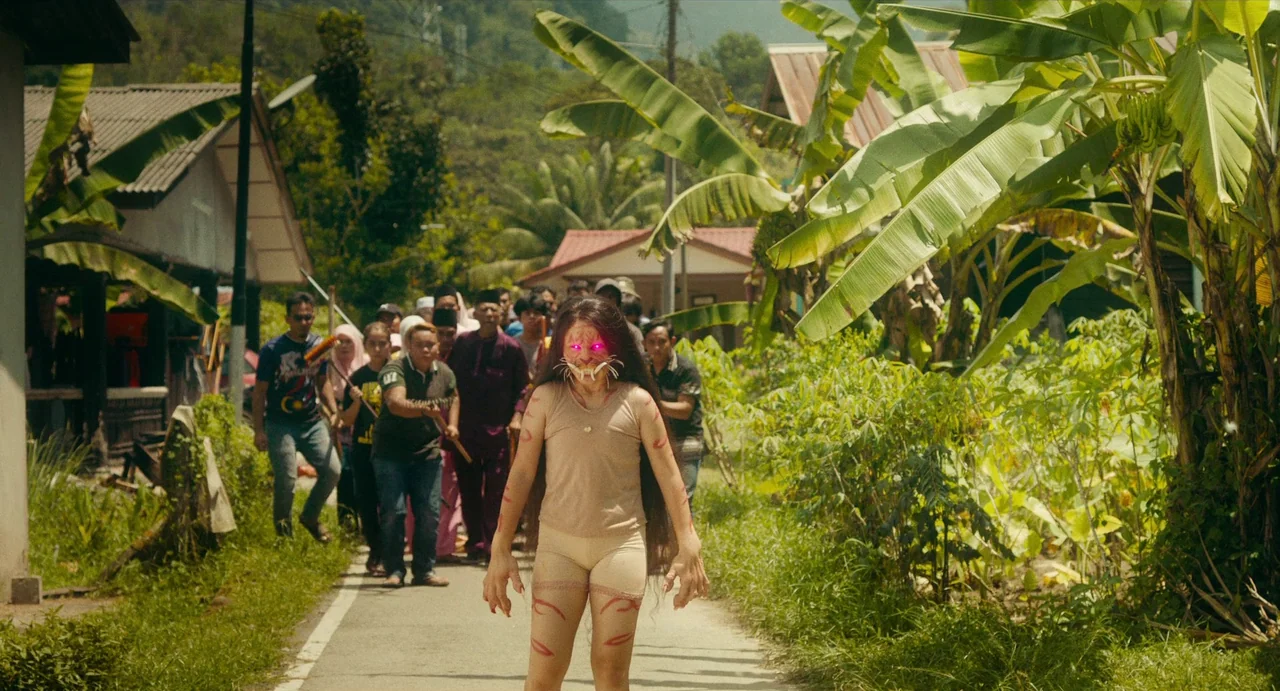 Tiger Stripes (2023) movie still: fullbody — A narrow paved path winds between rural village houses and lush green banana plants. A young woman with…