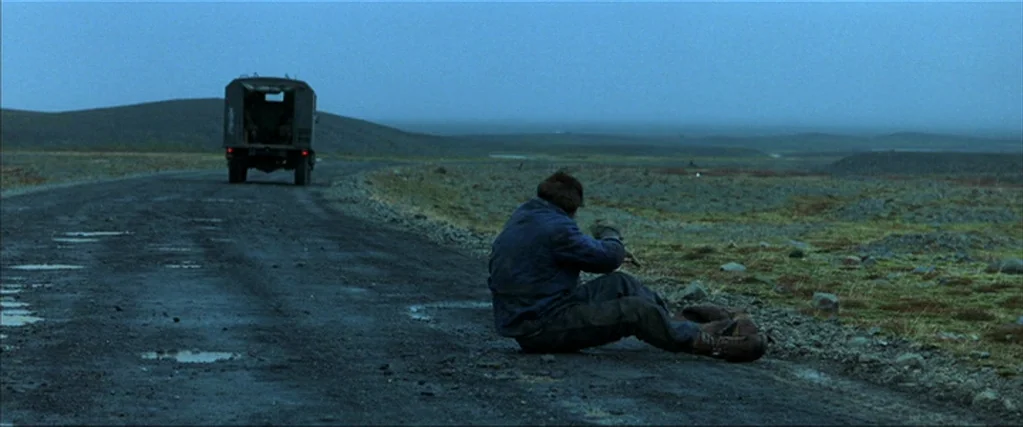 Batman Begins (2005) movie still: wide — A desolate gravel road stretches through a vast, barren landscape under a dim sky. A man sits on the side…