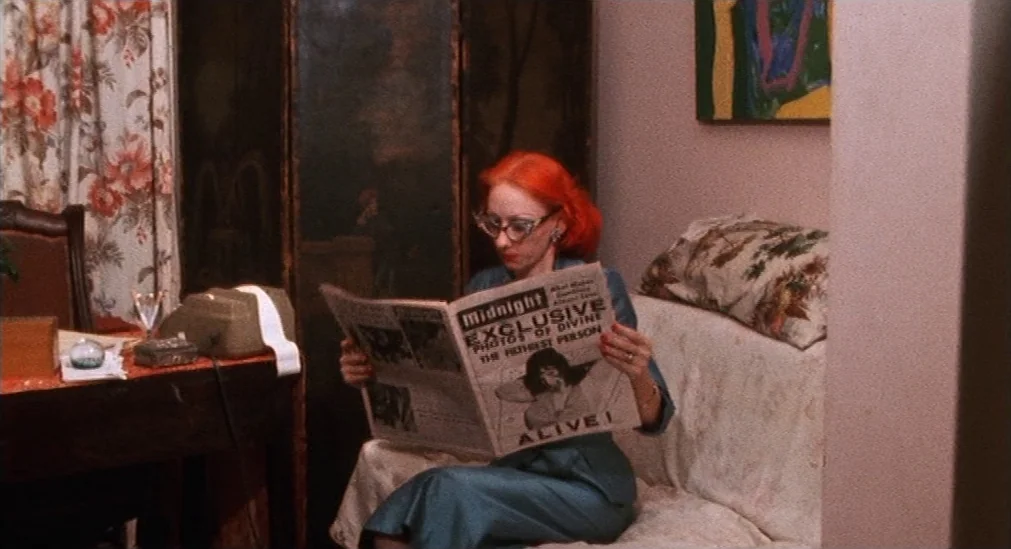 Pink Flamingos (1972) movie still: medium — A domestic space features a floral patterned sofa and a dark folding screen background. A woman sits on…