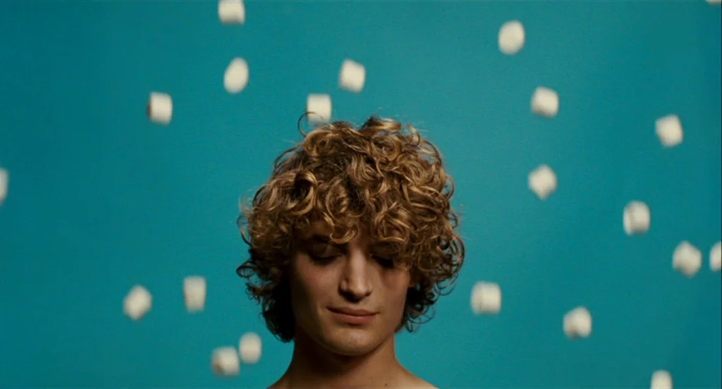 Heartbeats (Les Amours Imaginaires) (2010) movie still: closeup — A man stands in front of a flat turquoise studio backdrop. Multiple white…