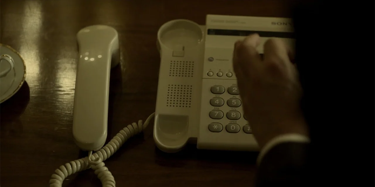 Oslo (2021) movie still: closeup — A dark wooden surface holds a white desk phone and a small tea saucer. A person's hand reaches in from the right…