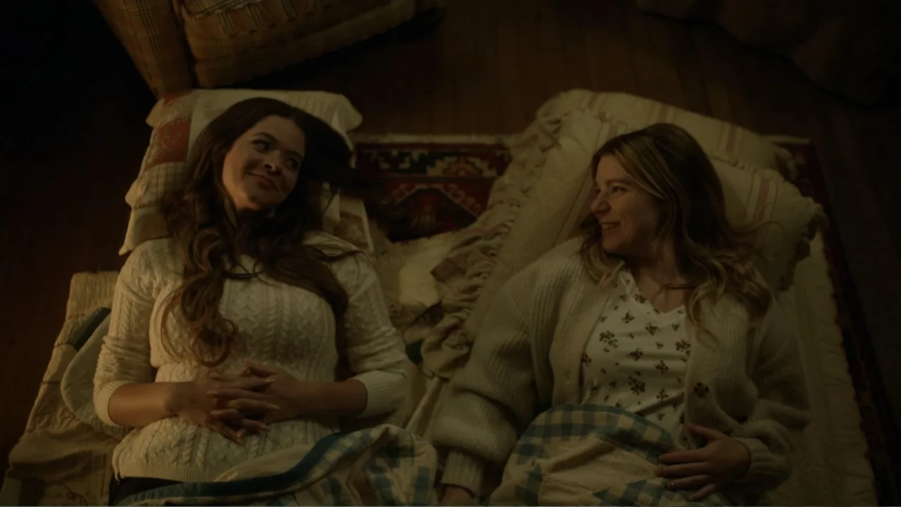 A Carpenter Christmas Romance (2024) movie still: medium — Two women lie on a blanketed floor in a dimly lit home setting. They face each other…