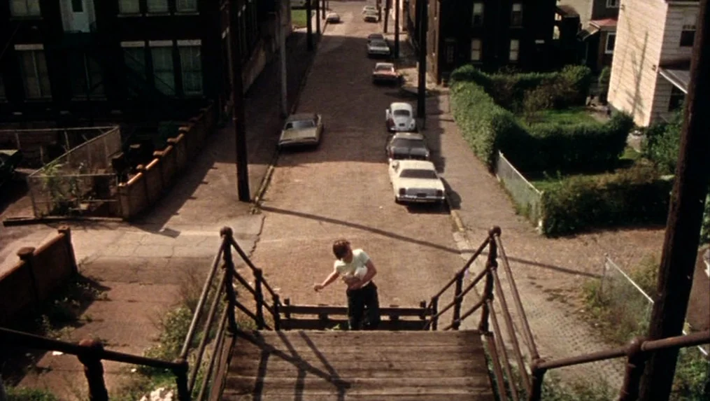 Martin (1977) movie still: wide — A residential city block features aging houses, parked vintage cars, and sparse green vegetation. A young boy…