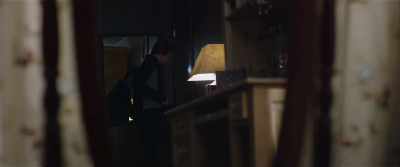 Captive (2020) movie still: medium — A dark bedroom interior features a desk and a lamp reflected in a mirror. A woman in a hoodie stands by the…