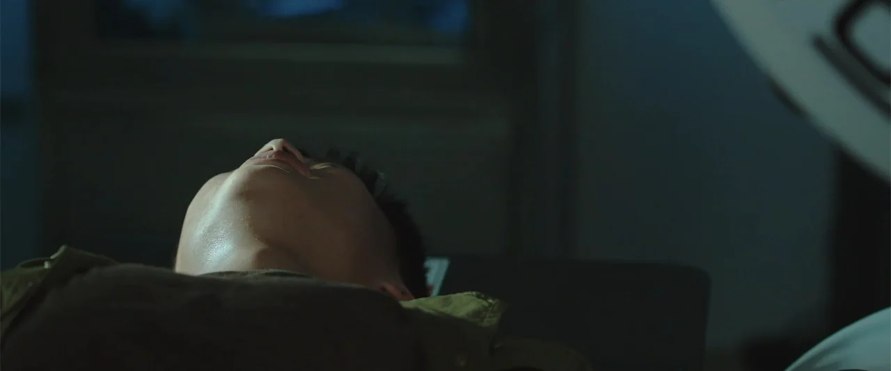 Presagios (2024) movie still: closeup — A dimly lit room contains a medical examination area with blurred architectural elements. A young man lies…