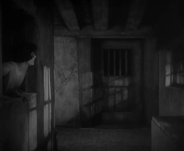 Sunrise (1927) movie still: medium — A dark stone passageway features heavy ceiling beams and a small barred window opening. A woman stands…