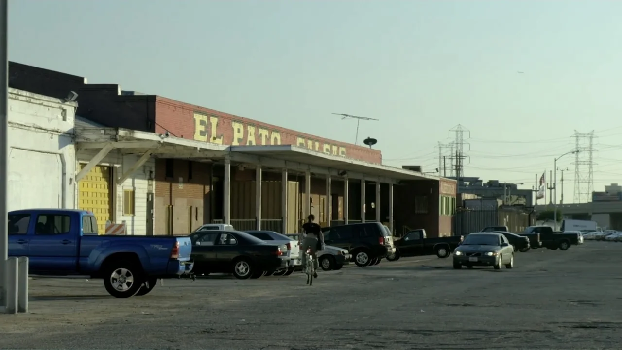 Loco Love (2003) movie still: wide — A vast, dusty parking lot sits before a low, red-brick commercial building with large lettering. A person rides…