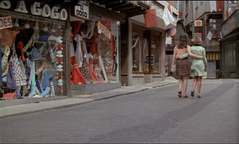 Small Change (1976) movie still: wide — A narrow European town street is lined with shop windows displaying colorful clothing merchandise. Two young…
