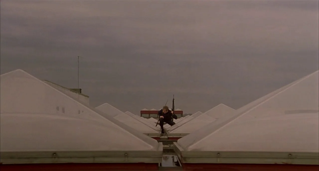 Dawn of the Dead (1978) movie still: wide — A flat industrial roof is covered in rows of triangular skylights under a cloudy sky. A man carrying a…