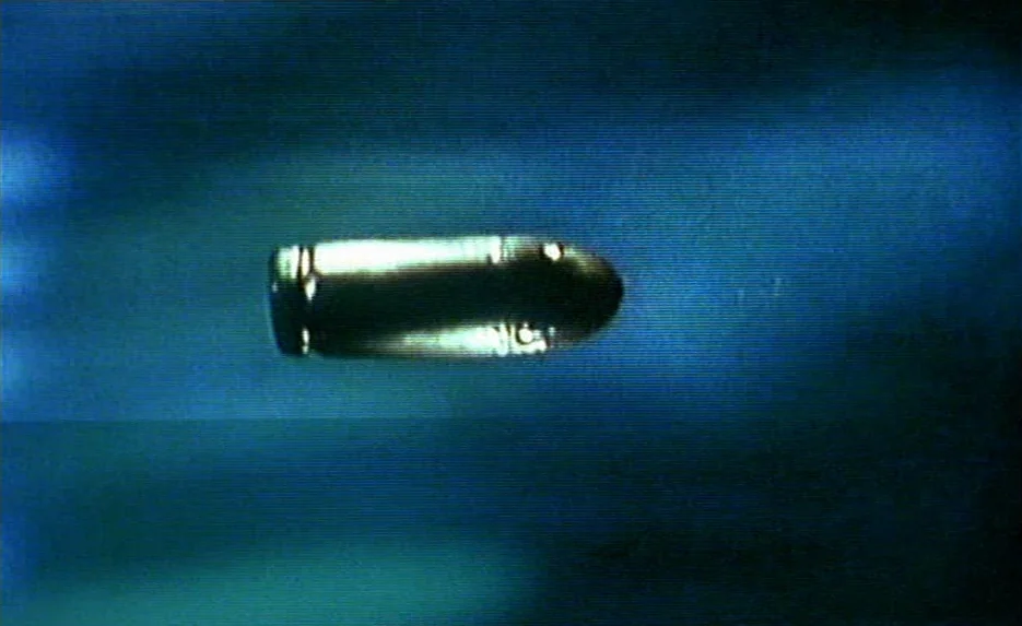 Irma Vep (1996) movie still: closeup — A brass bullet casing travels through dark blue void space. The image displays visible analog scan lines and…