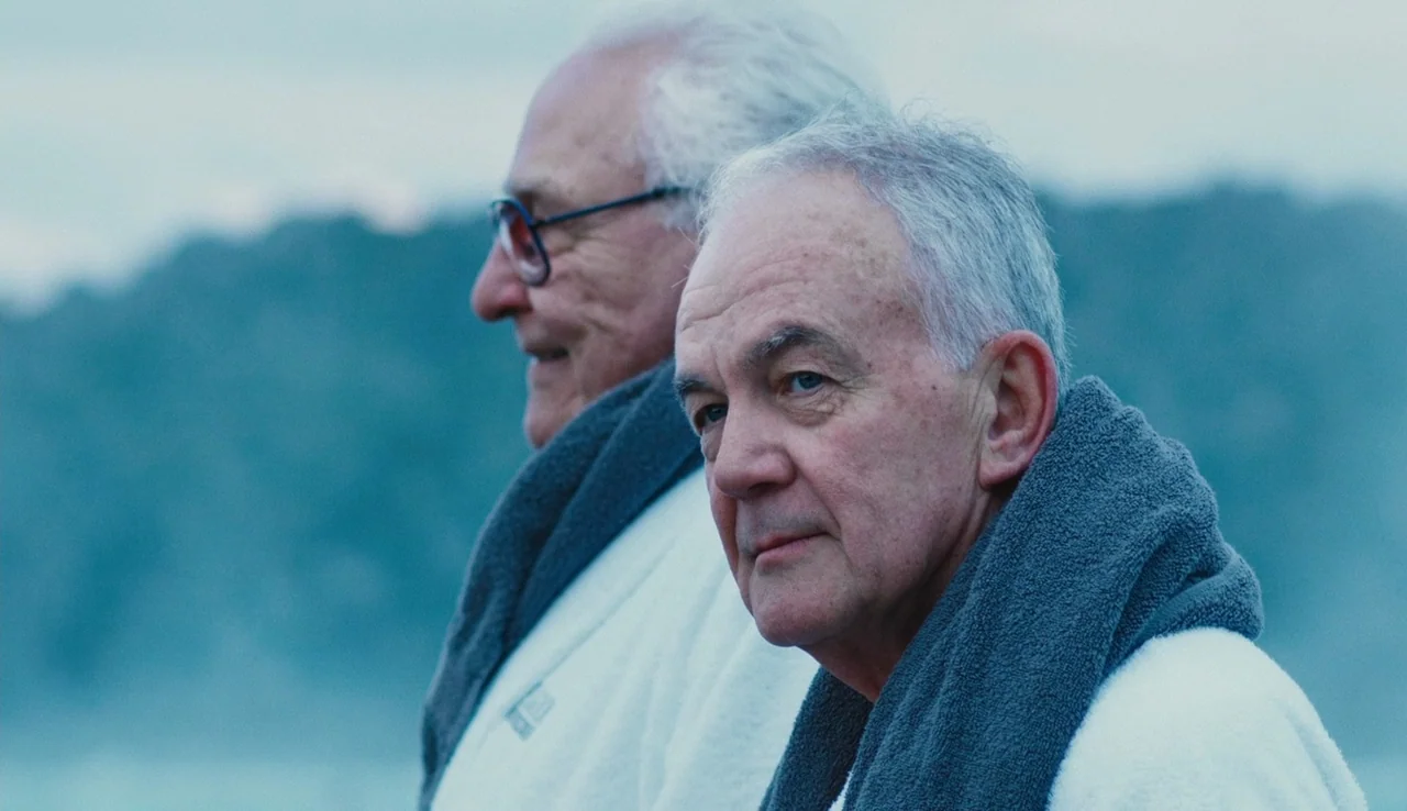 Land Ho! (2014) movie still: medium — Two elderly men in white bathrobes stand against a blurred, misty, cool-toned coastal background. Both men…