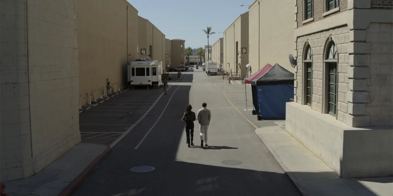A Hollywood Christmas (2022) movie still: wide — A paved narrow street flanked by tan industrial soundstage buildings on a film lot. Two people walk…