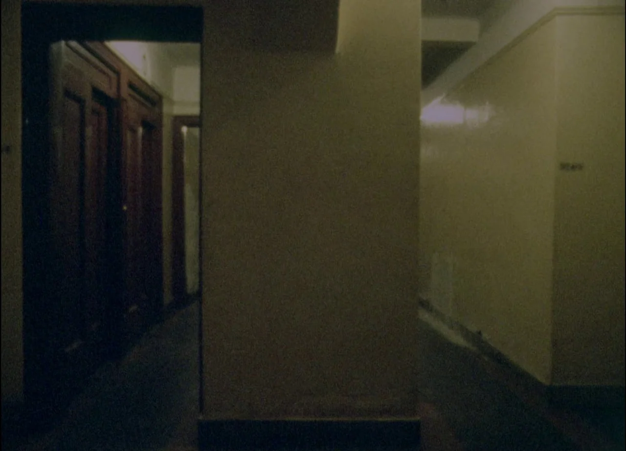 Hotel Monterey (1973) movie still: establishing — A dimly lit residential apartment hallway features dark wooden doors and pale yellow walls. An…