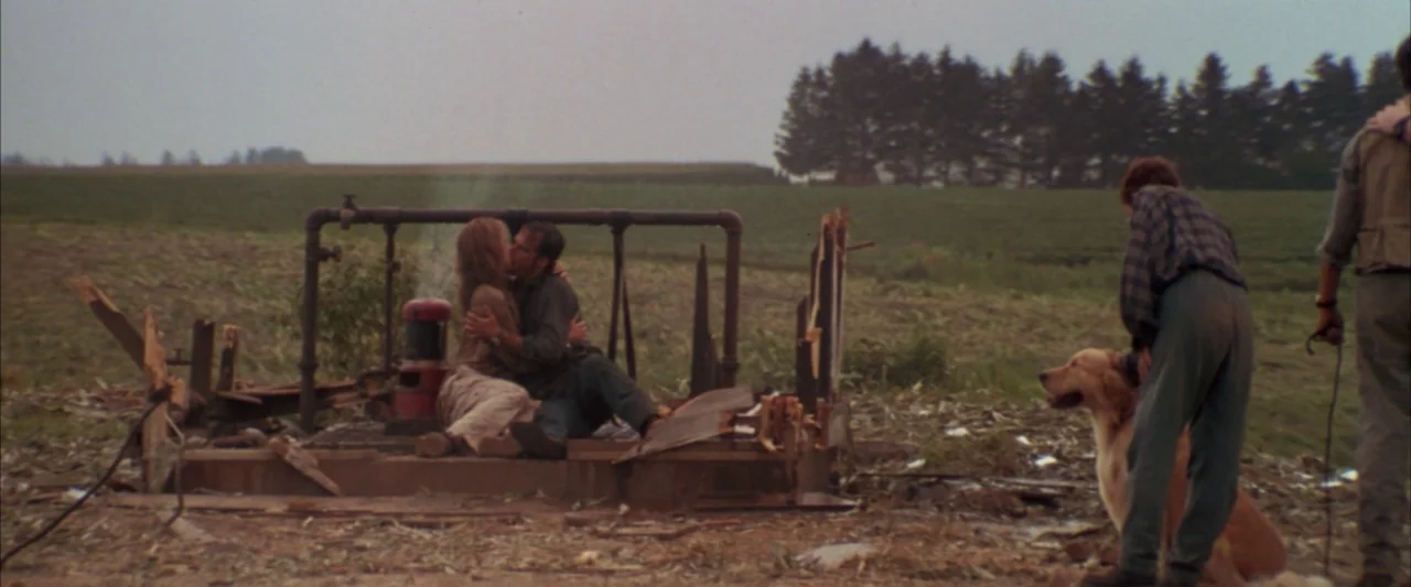 Twister (1996) movie still: wide — A desolate rural field contains a rusted, damaged industrial pump structure. A man and woman embrace on the…