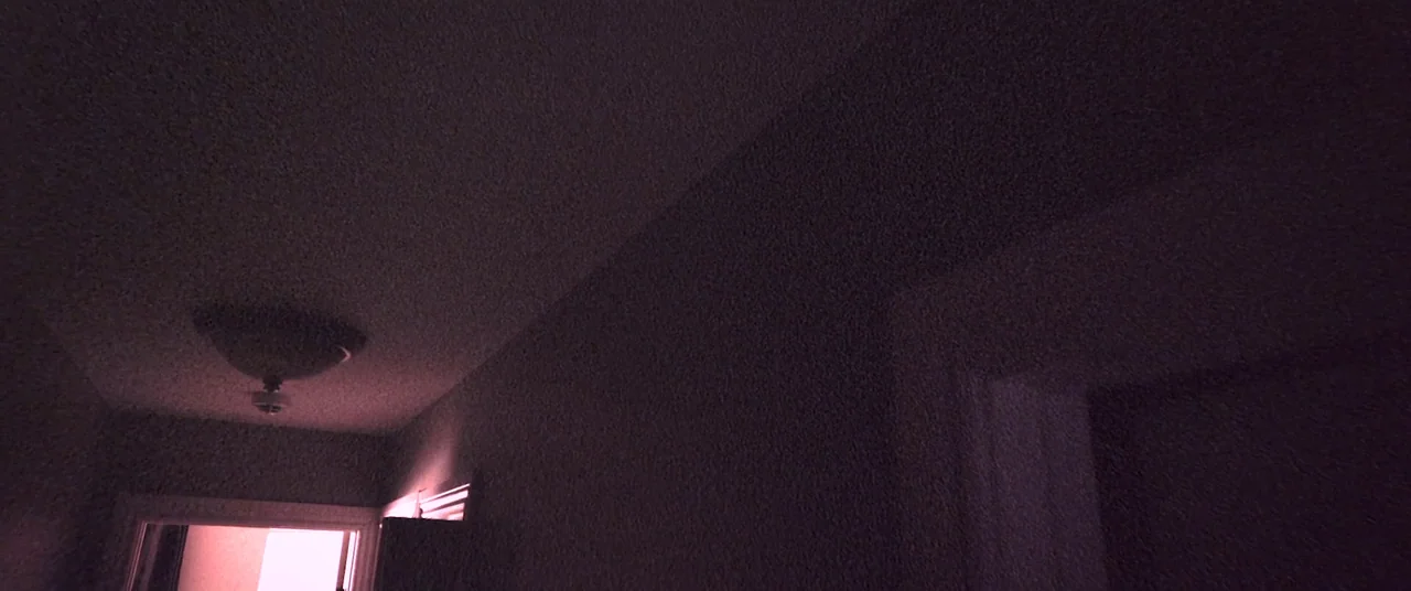 Skinamarink (2022) movie still: establishing — A dark, textured interior hallway shows the junction of a ceiling and wall. A small ceiling light…