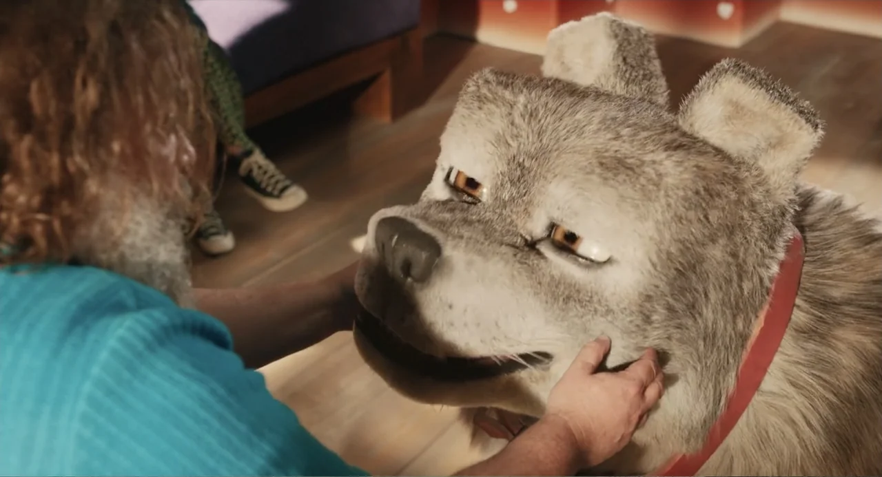A Minecraft Movie (2025) movie still: medium — An interior living room with wooden flooring and a person sitting near a puppet. A man in a blue…