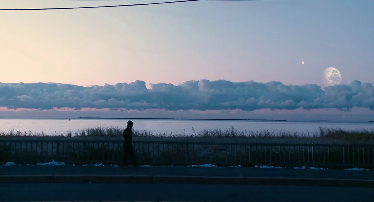 Another Earth (2011) movie still: wide — A dark, silhouetted figure walks along a road bordering a calm body of water. Two planetary bodies are…