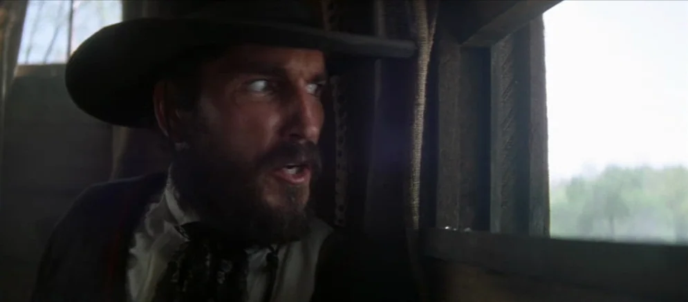 Ride With The Devil movie still: closeup — An interior view of a wooden wagon with a small rectangular window cut out. A man with a dark beard…