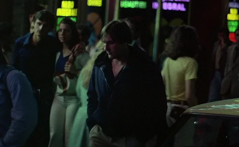 Maîtresse (1975) movie still: medium — A night street scene features a man walking past a series of illuminated storefronts. Several other people…