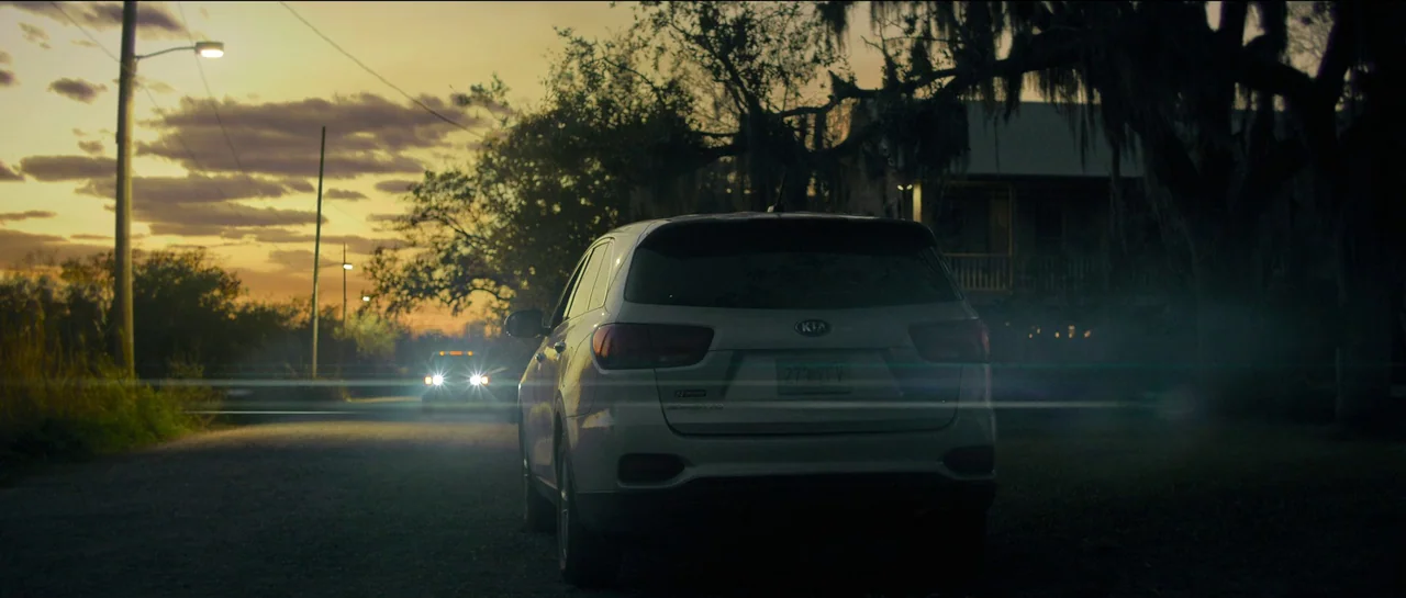 The Killer (2023) movie still: wide — A white SUV is parked on a dirt shoulder during a golden hour sunset. A second vehicle approaches from the…