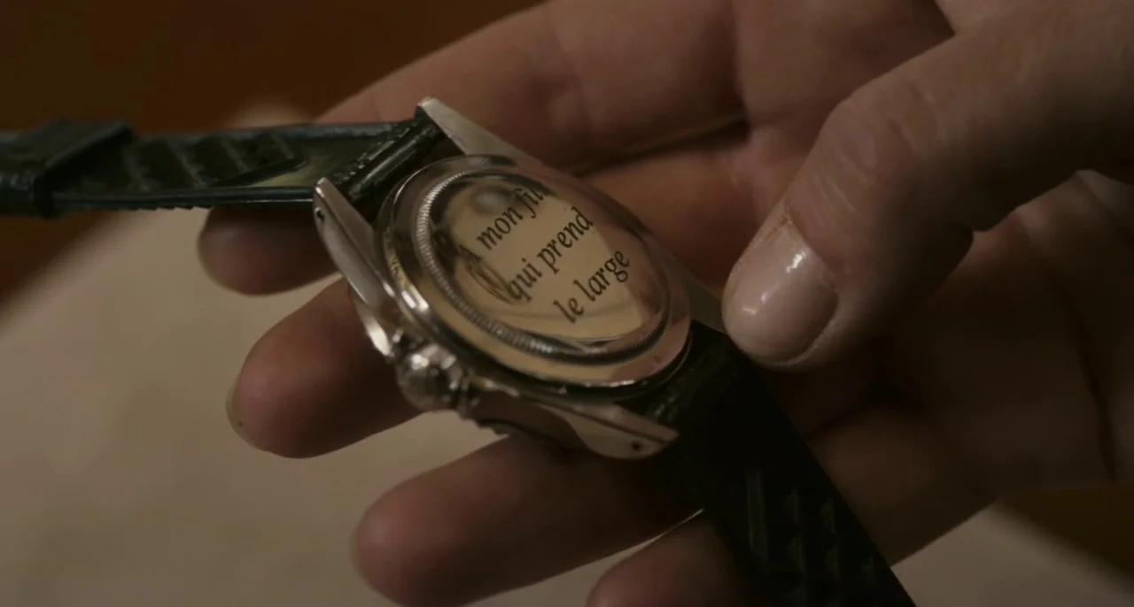 Bastards (2013) movie still: closeup — A close-up view shows a hand holding a silver watch with a leather strap. The back of the watch case features…