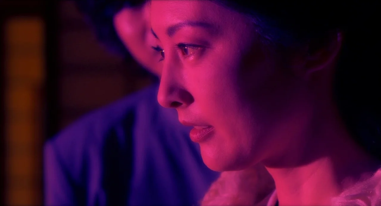 Hanagatami movie still: closeup — A dark indoor space is filled with intense pink and purple colored light. A woman in the foreground looks forward…