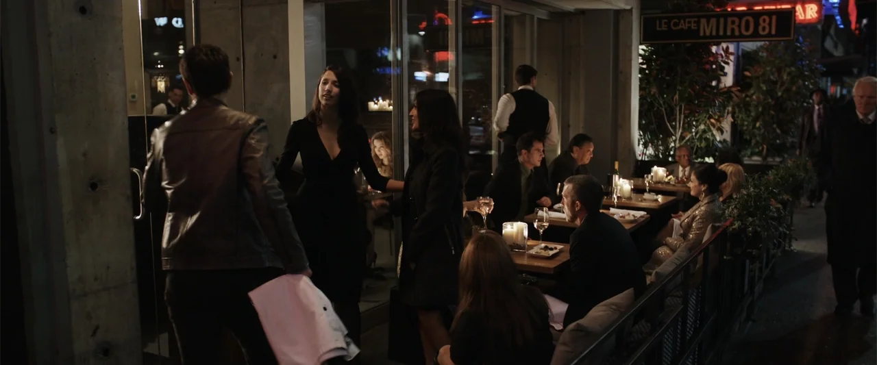 Final Destination 5 (2011) movie still: medium — A bustling cafe terrace at night with glass doors and wooden tables with candles. Two women stand…