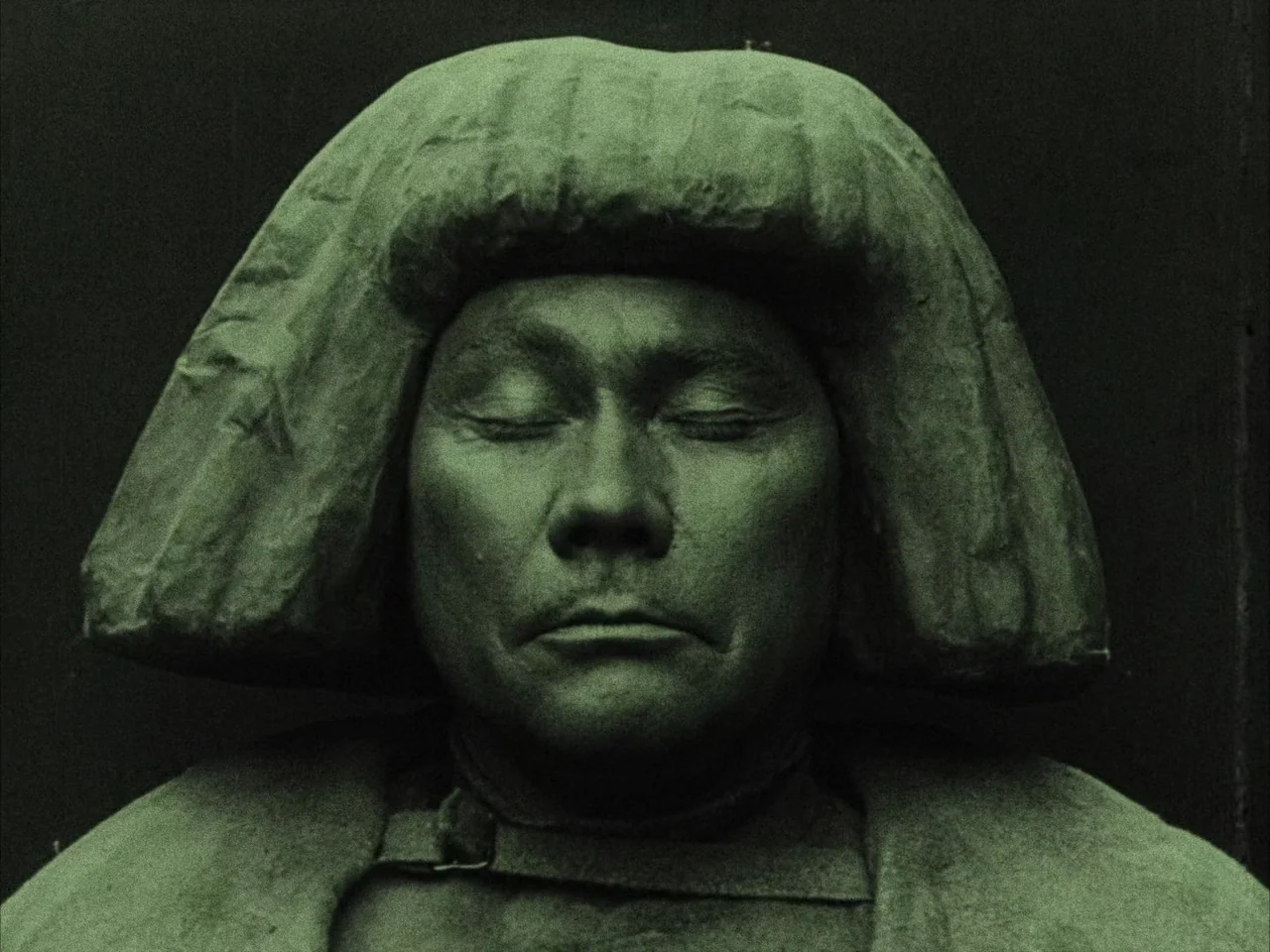 The Golem (1920) movie still: closeup — A dark stone sculpture of a man rests against a matte black wall background. The figure has eyes closed and…