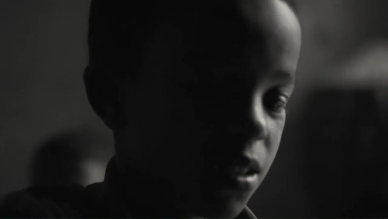 Faya Dayi (2021) movie still: closeup — A dark interior space with minimal light cast upon the subject. A young boy looks down with his features…