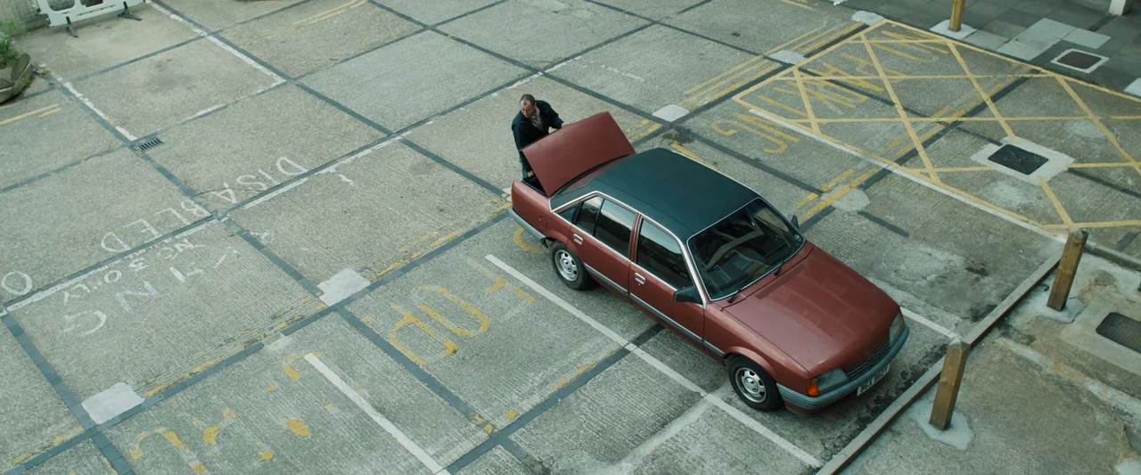 Black Sea (2014) movie still: wide — A concrete parking lot features faded yellow painted markings on the weathered pavement surface. A man in dark…