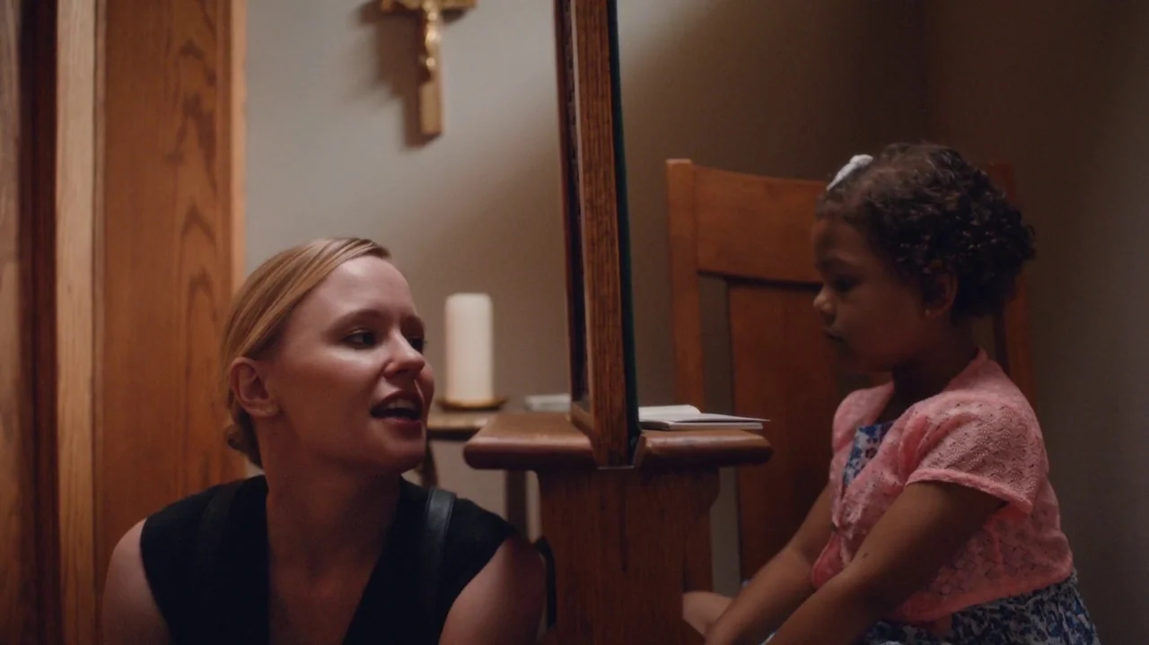 Saint Frances (2019) movie still: medium — A quiet indoor space features a wooden kneeling bench and a wall-mounted crucifix. A woman in a black…